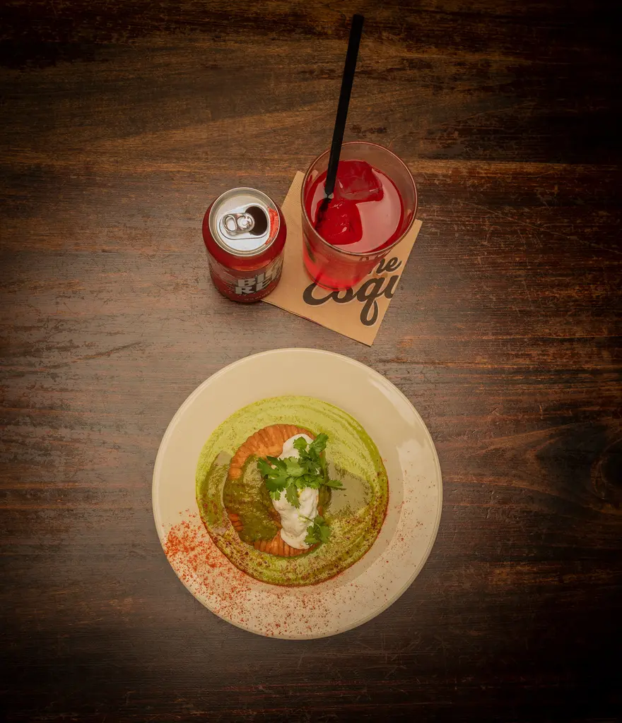 An overhead view of a plated dish with green sauce, a can of Big Red and a red drink on a wooden table.