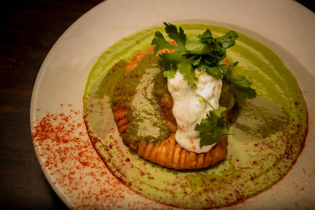 A golden pastry under green sauce, crema and cilantro on a cream-colored plate dusted with red spice.