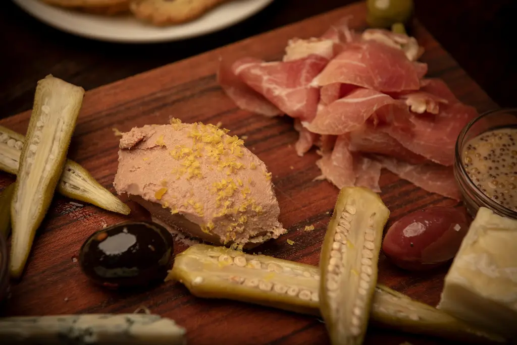A close view of pâté topped with lemon zest beside prosciutto, olives and pickled okra on a wooden board.