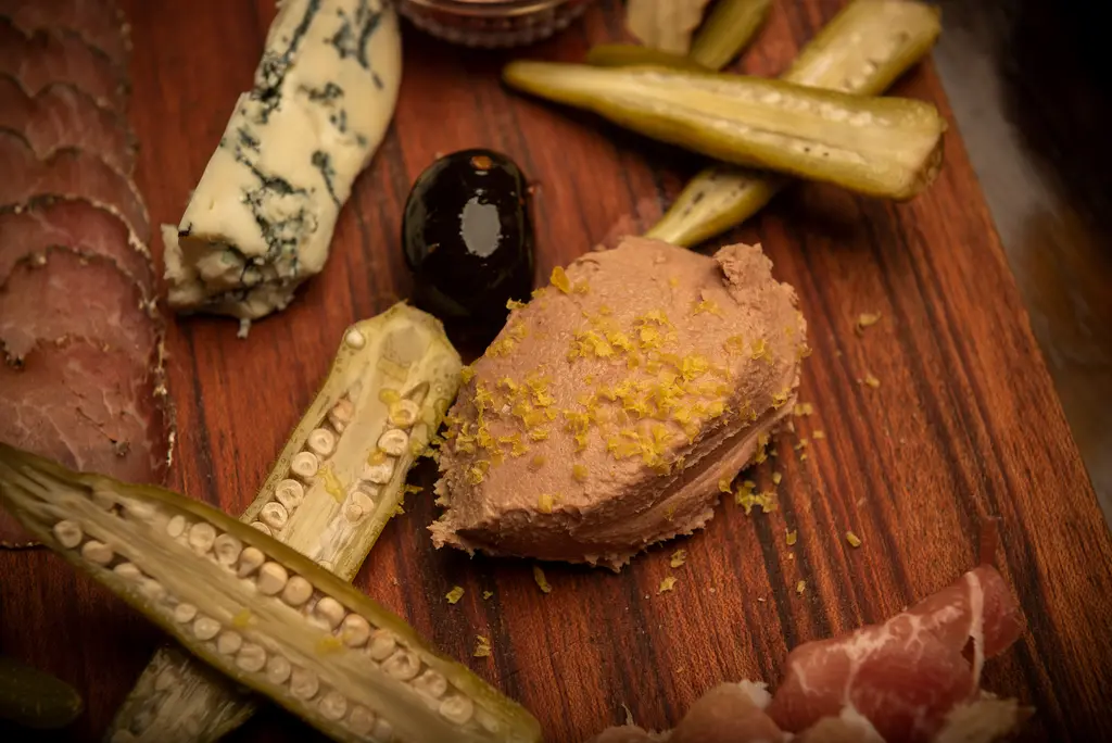 A close view of pâté dusted with lemon zest beside a black olive, blue cheese and pickled okra.