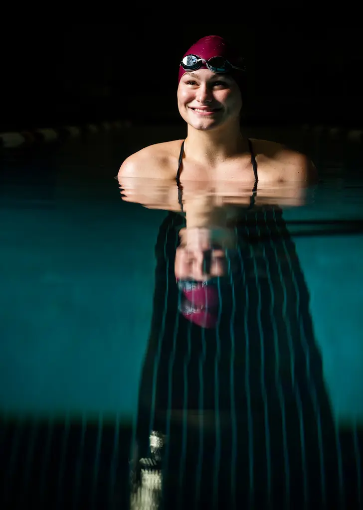 A smiling swimmer in a maroon cap with goggles stands in a dark pool, her reflection stretching across the water below.