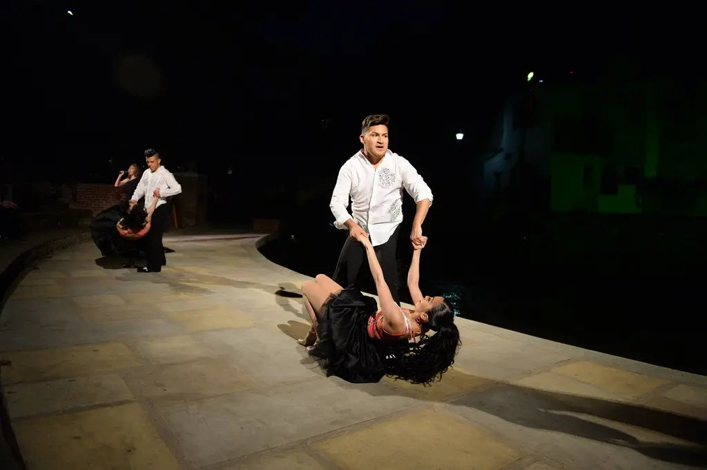 A man in a white shirt holds a woman in a black ruffled dress in a low dip on a lit stone walkway at night, with another couple behind them.