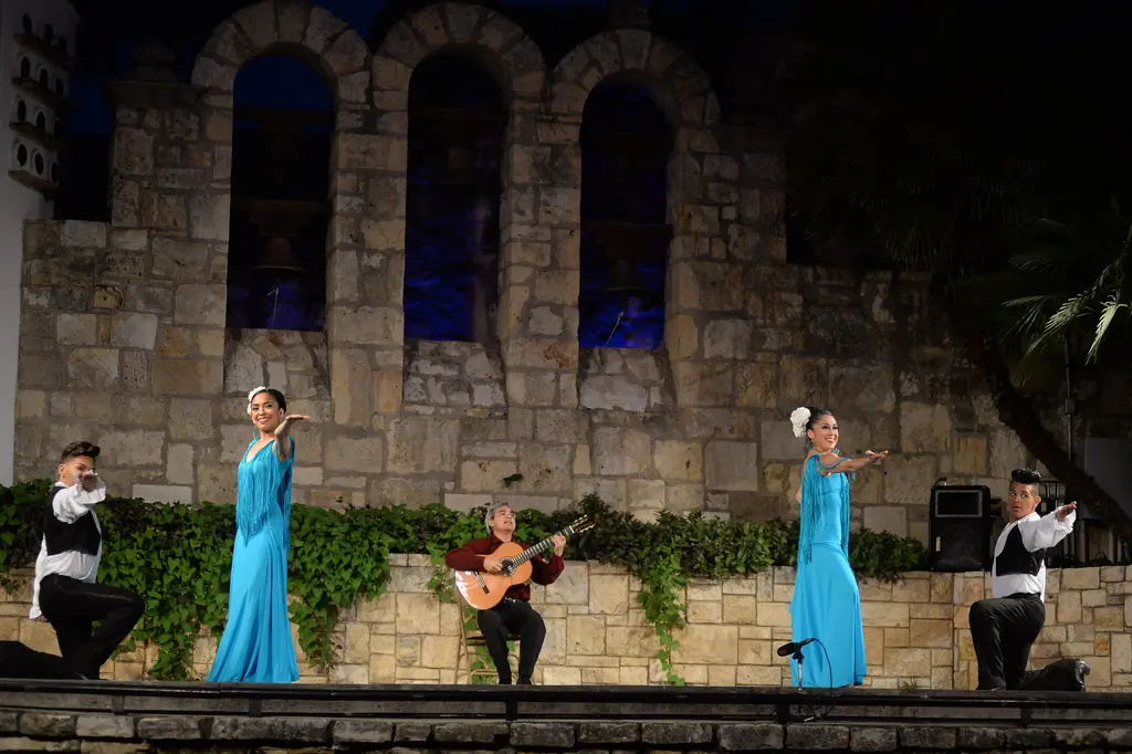 On the stone stage, two women in long turquoise dresses and two kneeling men in black vests surround a seated guitarist.