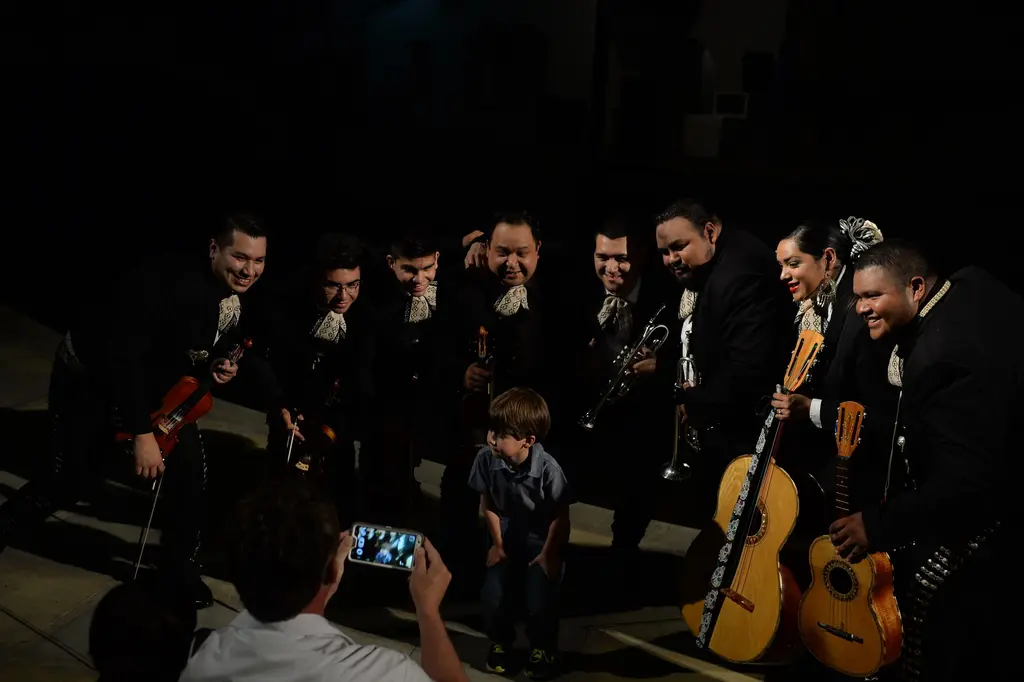 A small boy stands in front of a smiling mariachi band holding violins, a trumpet and guitars as a man takes his picture with a phone.