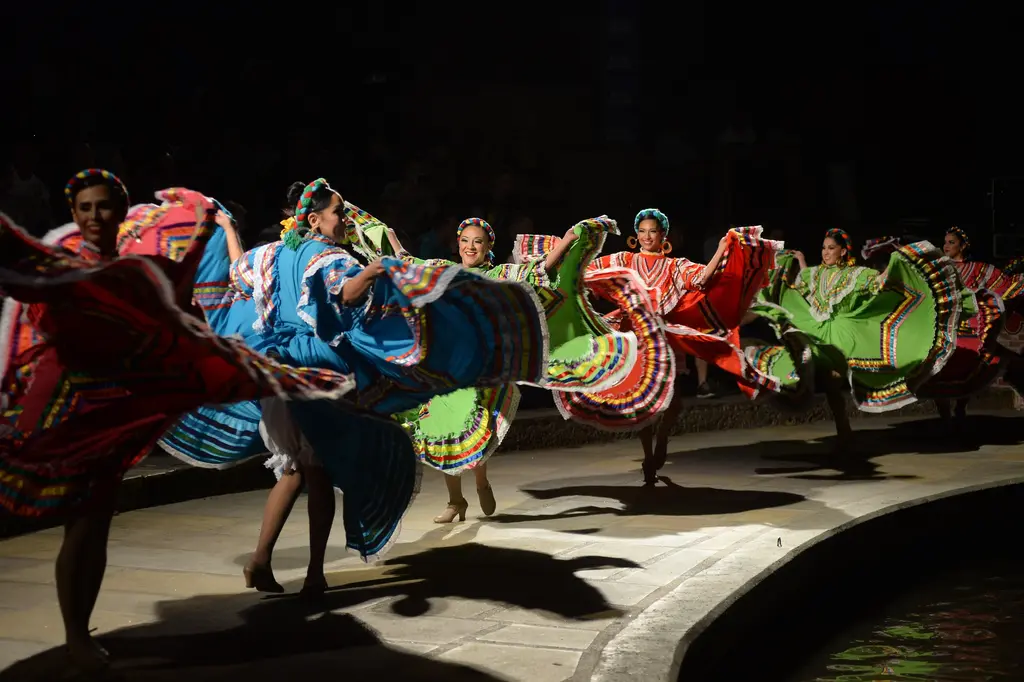 A line of dancers in blue, red and green dresses spins along the curved riverside walkway at night, their shadows on the stone.