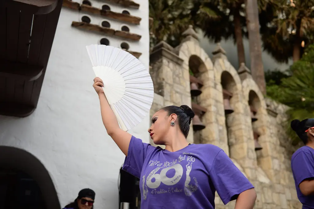 A dancer raises a white fan above her head in front of a stone wall with arched openings that hold bells.