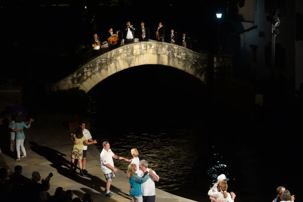 Couples dance on the stone riverbank at night while a mariachi band plays on the arched footbridge above them.