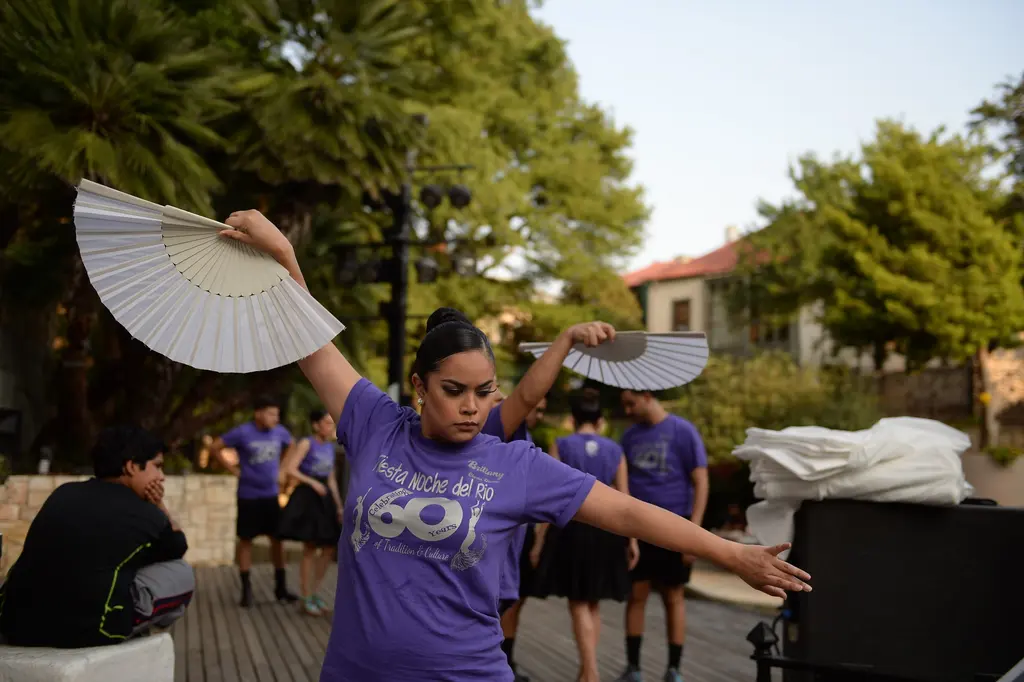 A dancer in a purple T-shirt stretches one arm out and holds a white folding fan overhead, with other dancers behind her under the trees.