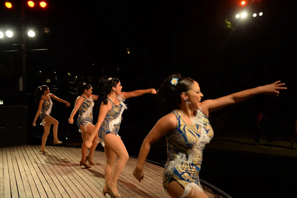 Four dancers in sparkling blue and white costumes with feather trim stride across a wooden stage under bright stage lights.