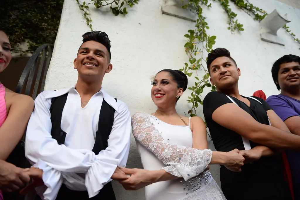 A young man in a white shirt and black vest stands hand in hand with a woman in a white lace dress and other dancers against a vine-covered wall.