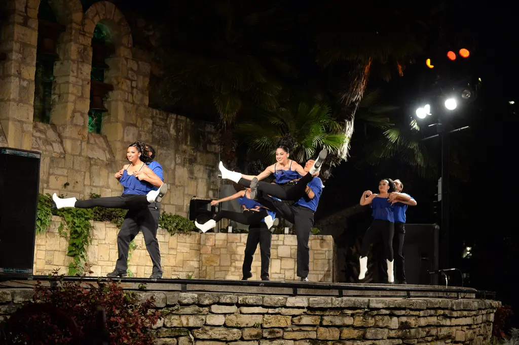 Pairs of dancers in blue shirts and black pants kick high on the stone stage at night.