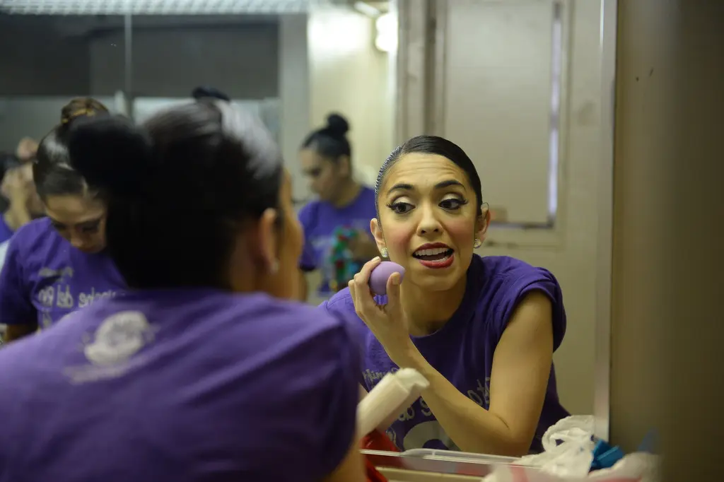 A dancer in a purple shirt holds a makeup sponge to her cheek and talks while looking into a mirror.