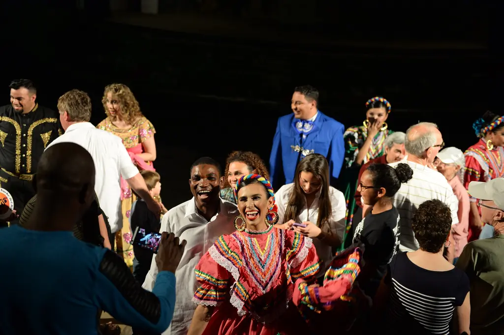 A smiling dancer in a ruffled folklorico dress poses with audience members holding up phones as other performers mingle in the crowd.