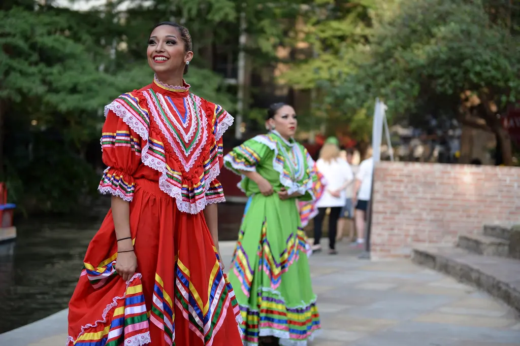A smiling dancer in a red ruffled dress with ribbon stripes stands by the river, with a dancer in a green dress behind her.