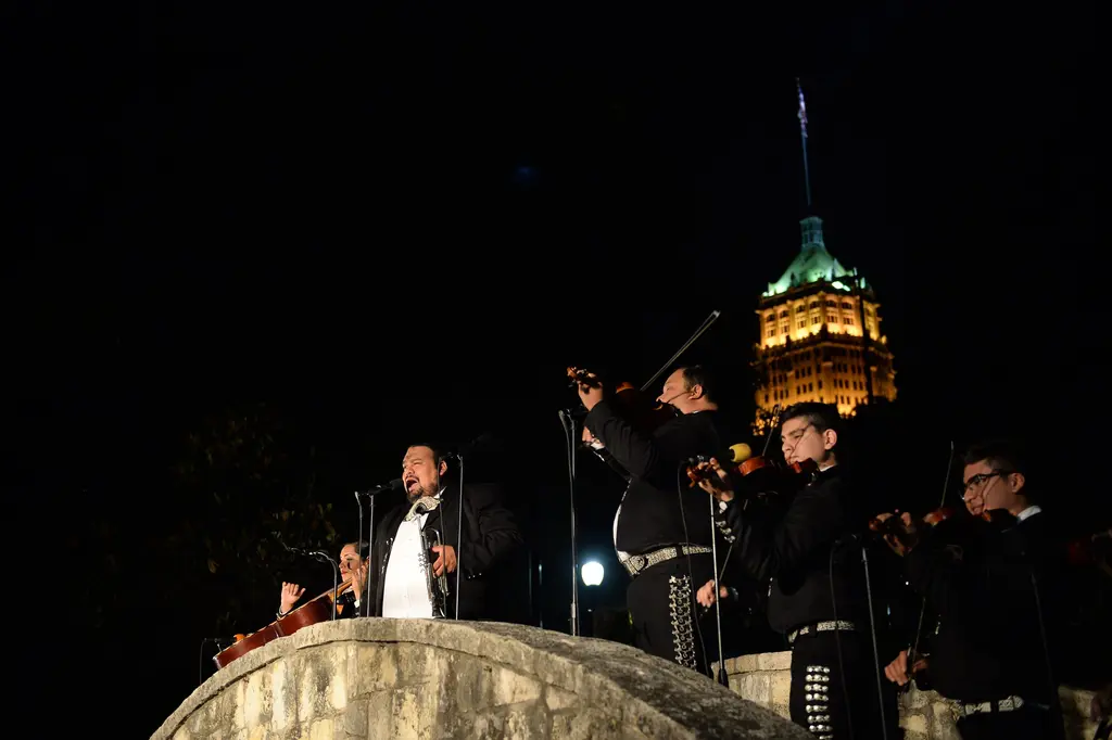 A mariachi singer in a black suit sings into a microphone on the arched stone bridge, with violinists beside him and a lit tower behind.