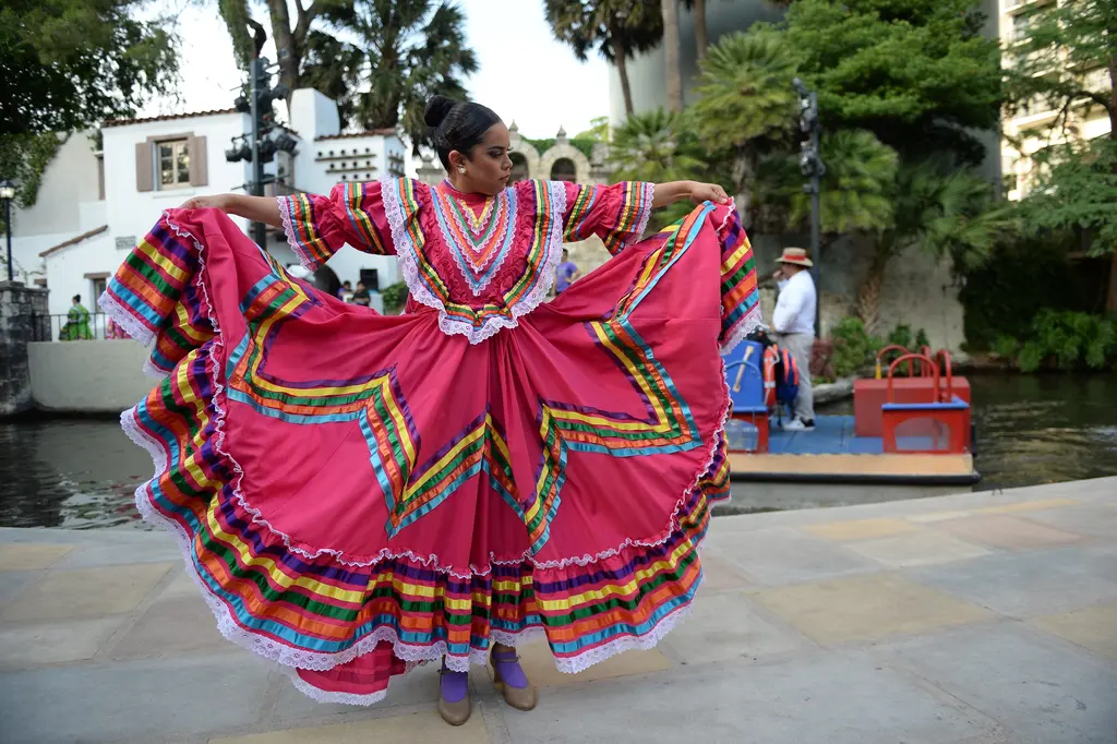 A dancer in a bright pink dress with rainbow ribbon trim holds her wide skirt out at both sides beside the river.