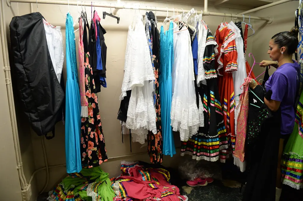 A dancer reaches for a dress on a packed clothing rack of colorful ruffled folklorico dresses and white lace gowns.