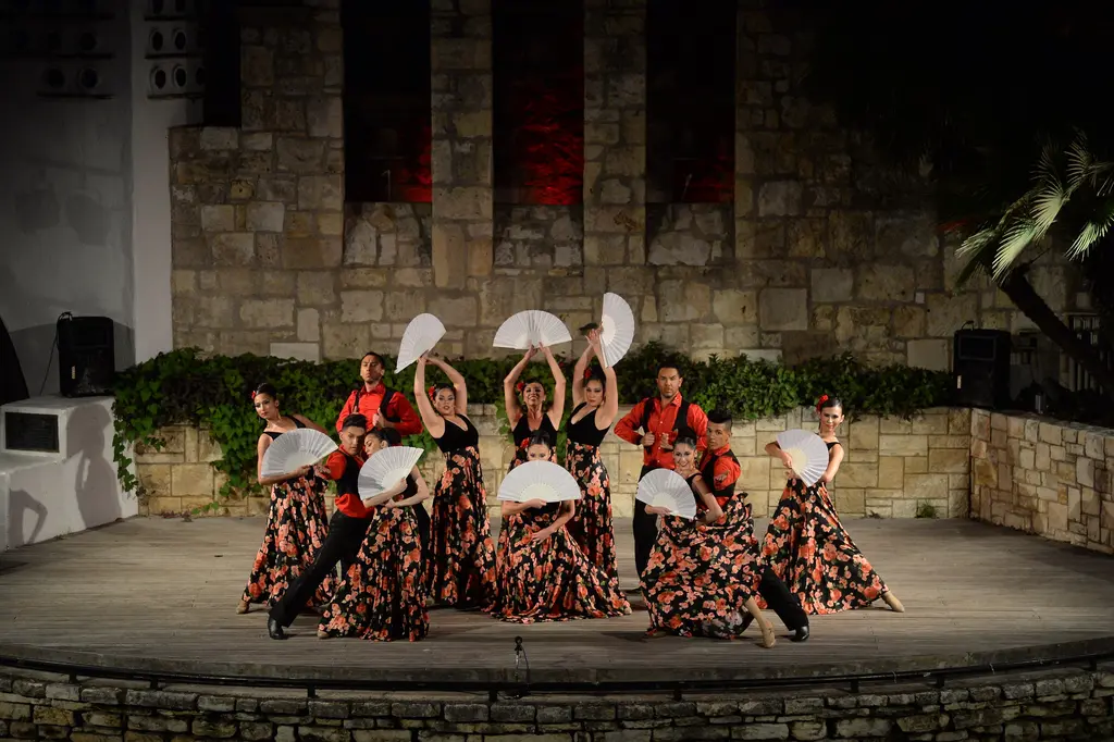 On the lit stage, women in floral skirts holding white fans pose with men in red shirts in front of the stone wall.