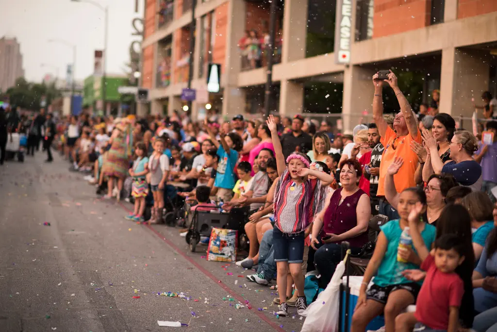 Spectators seated along a street lined with buildings wave and cheer; a girl in a pink flower crown raises one arm while a man in an orange shirt holds up a phone, with confetti in the air.