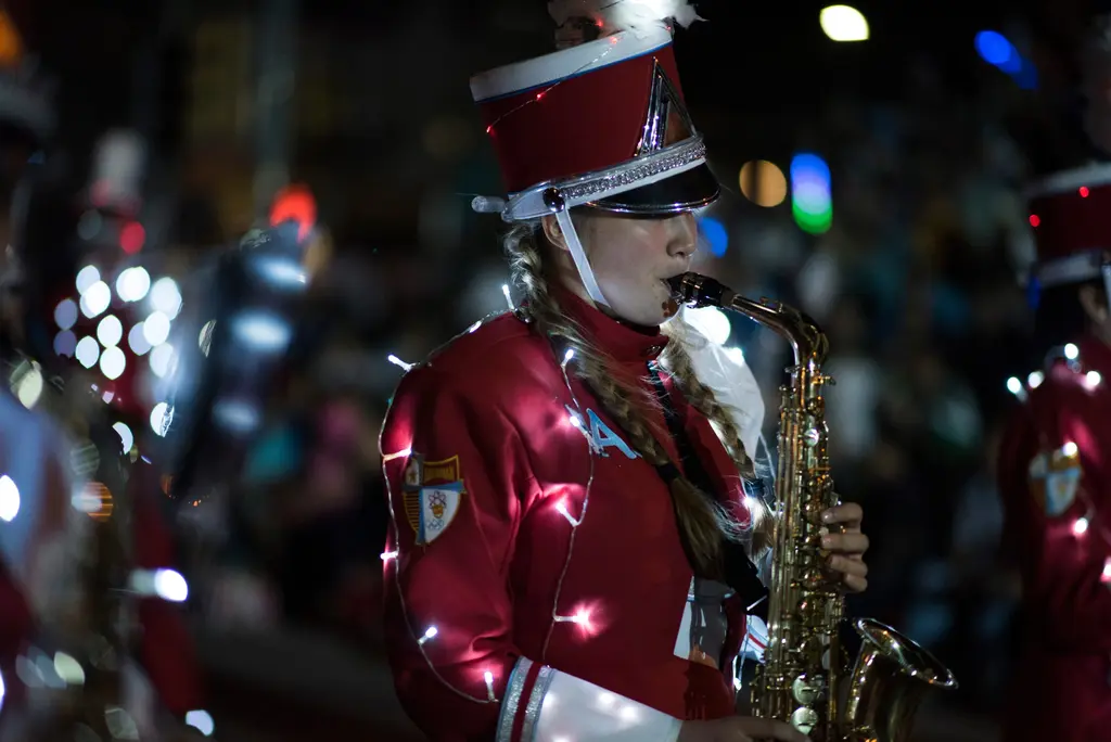 A young woman with braided hair, a tall red-and-white shako and a red band jacket wrapped in small white lights plays an alto saxophone, with blurred colored lights behind her.