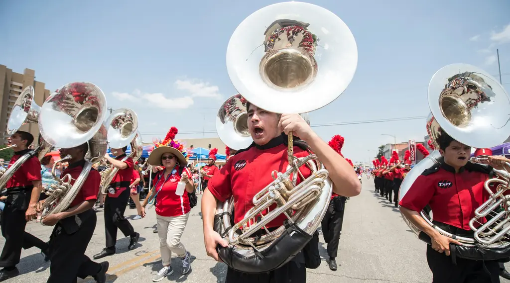 A young man in a red shirt and black pants carries a silver sousaphone with its bell high above him, with other sousaphone players in red and band members in red-plumed hats behind.