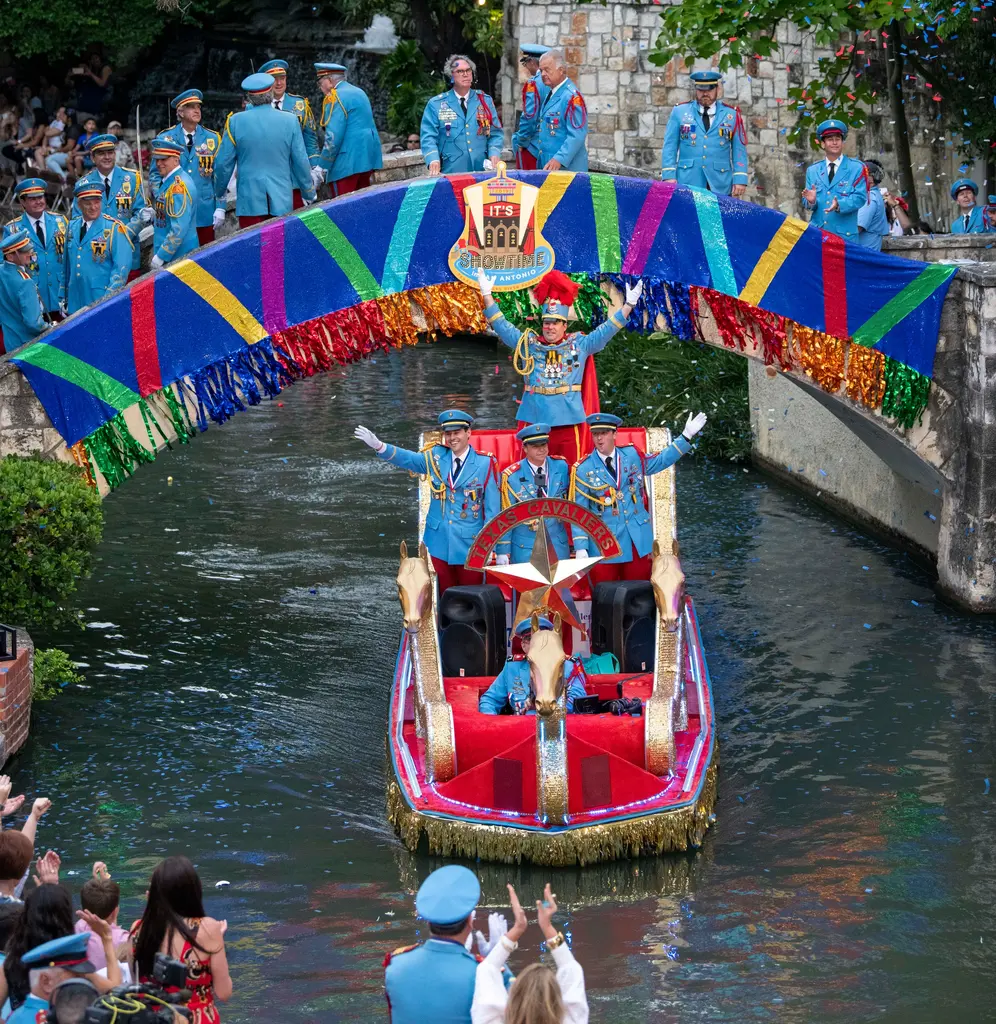 Seen from above, a red barge with gold trim carries men in light-blue uniforms under a sequined striped bridge where more uniformed men stand.