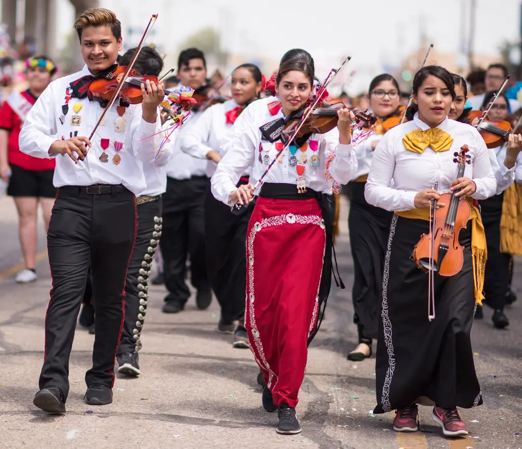 A young man and two young women in white shirts, bow ties and charro-style trousers and skirts play violins while walking, with medals pinned across their chests.
