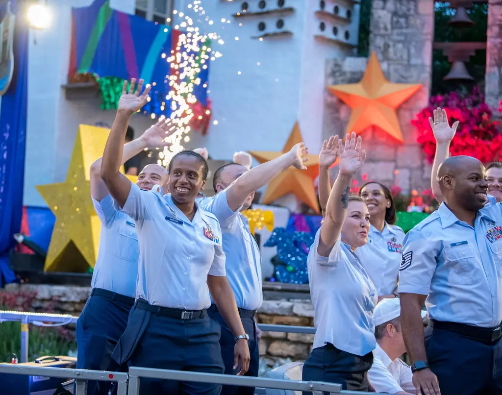 Smiling service members in light-blue short-sleeve uniform shirts wave with raised arms on a barge, with glittering gold and orange stars behind them.