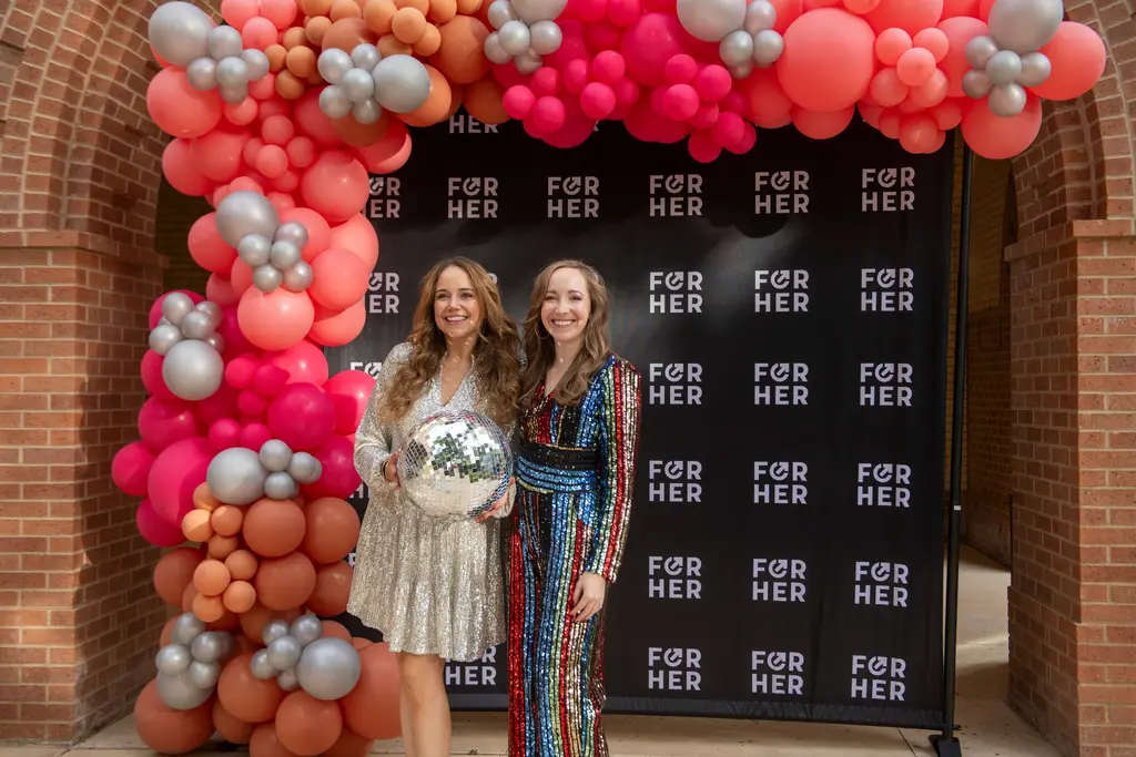 Two women in sequined outfits, one holding a disco ball, pose under a pink, coral and silver balloon arch at the For Her gala.