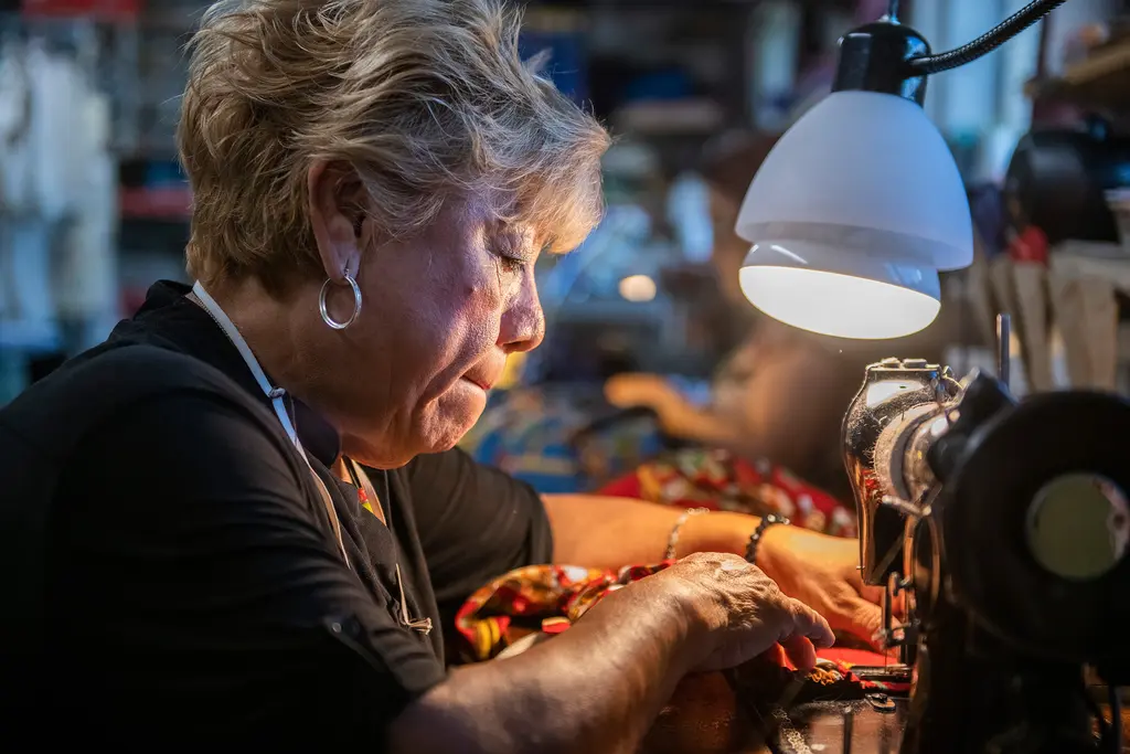 A close view of a woman with short blond hair guiding fabric through a sewing machine under a lamp.