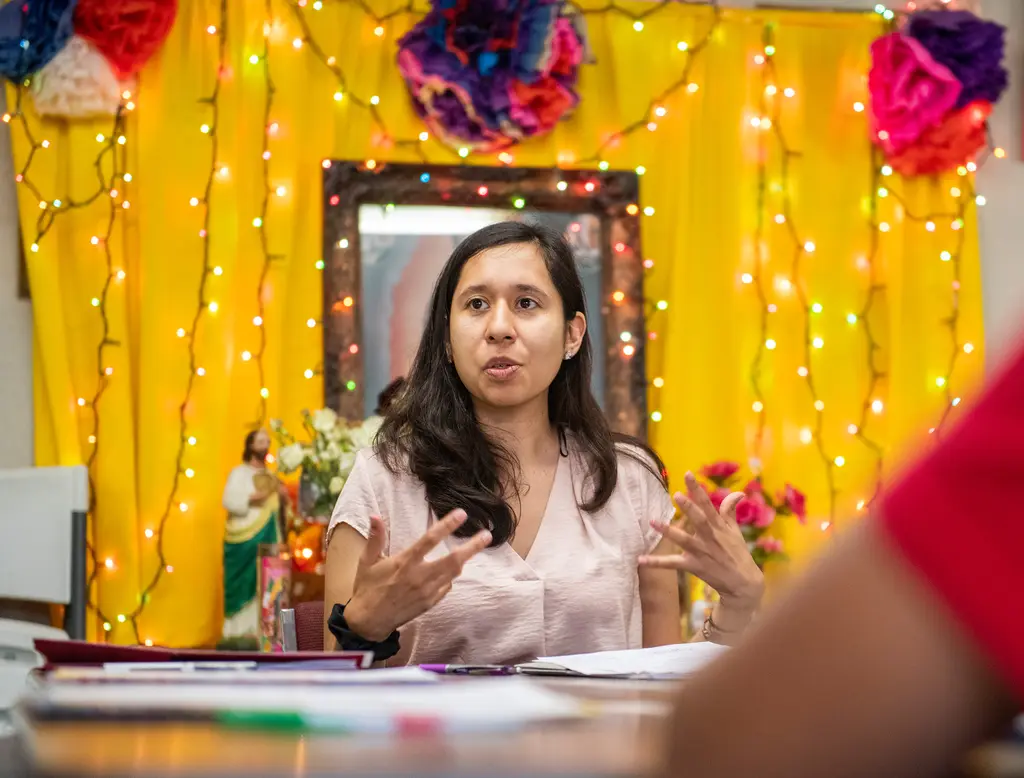 A young woman in a pink blouse gestures as she speaks at a table in front of a yellow altar hung with string lights and paper flowers.