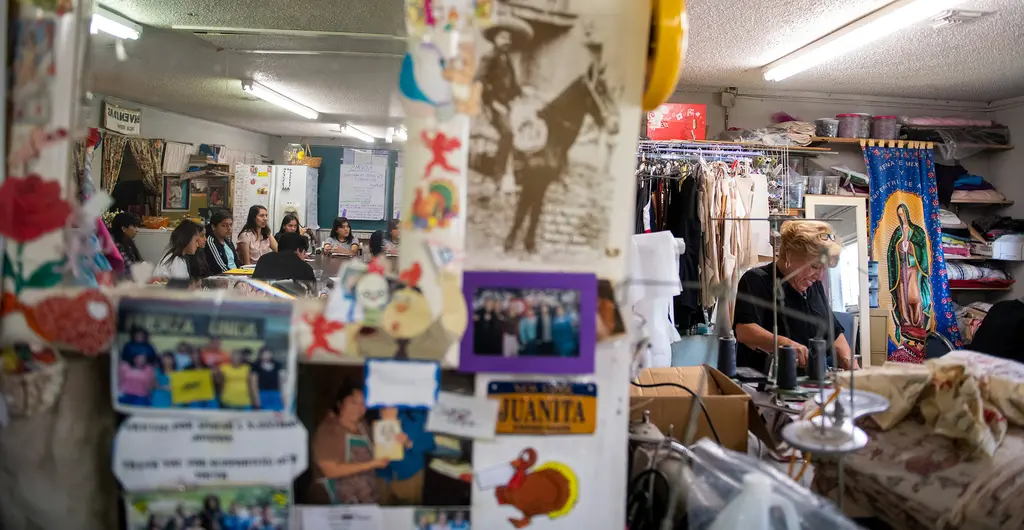 A wall covered in photos, flyers and mementos, with women working at sewing machines beyond it.