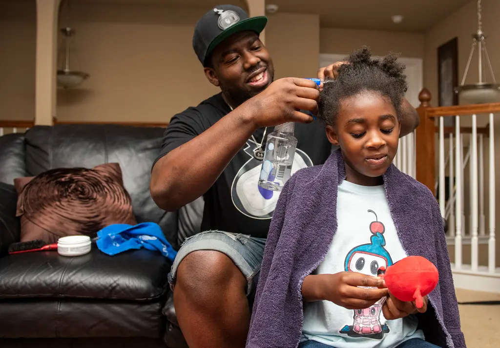 Gilbert Glaster, in a black cap and robot T-shirt, sprays and combs his daughter's hair as she sits on a couch holding a stuffed toy.