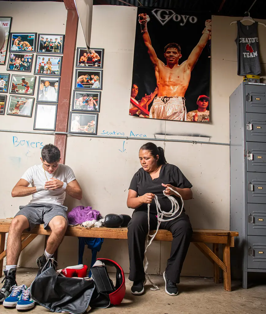 A young boxer wraps his hands as his mother untangles hand wraps on a bench beneath a large Goyo poster and framed fight photos.