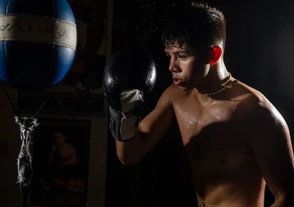 A sweating young boxer raises his glove beside a blue double-end bag in a dark gym.
