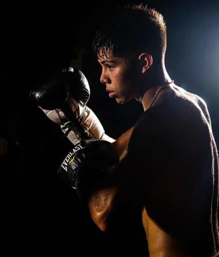 A sweating young boxer holds his gloves high in a dark gym, lit from behind.