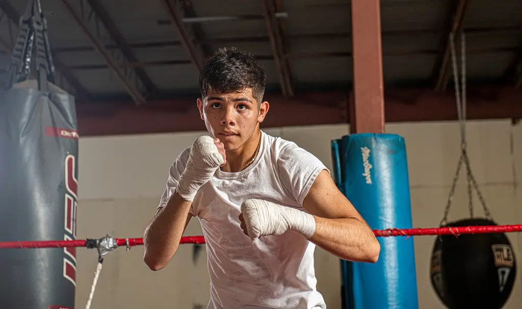 A young boxer in a white T-shirt and hand wraps takes his stance in the ring, heavy bags behind him.