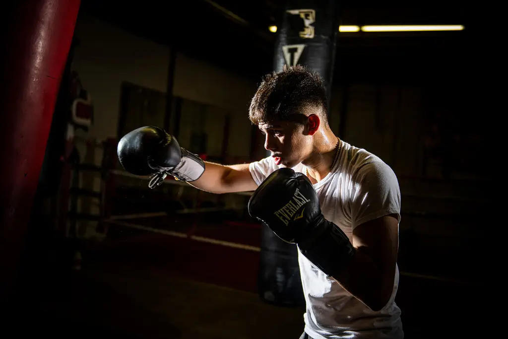 A young boxer in a white T-shirt throws a jab toward a heavy bag in a dark gym, lit from the side.