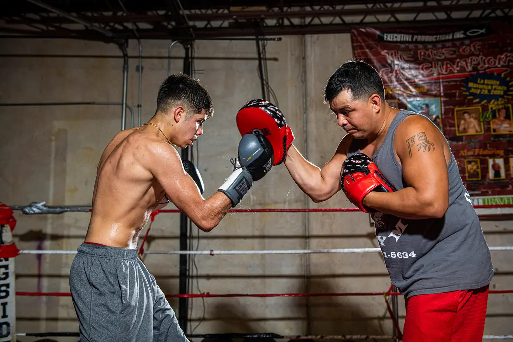 A shirtless young boxer throws a punch into a red mitt held by a trainer in a gray tank top inside the ring.