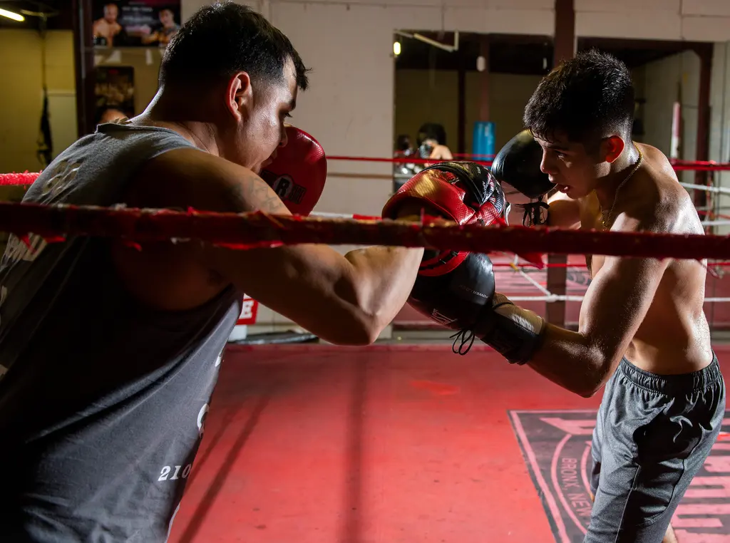 A young boxer throws a punch at a trainer's red mitt across the red ring ropes.