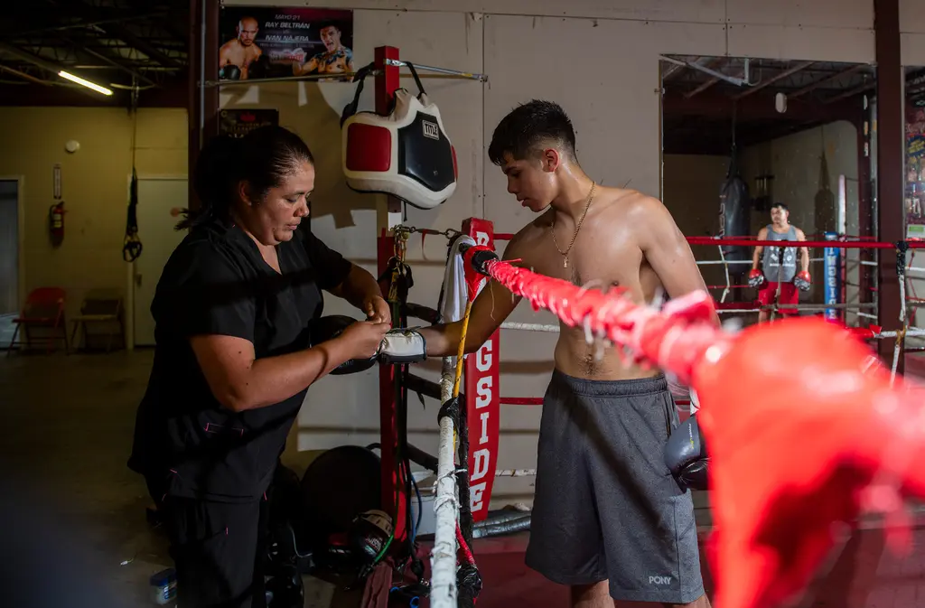 A woman ties a young boxer's glove at the corner of the ring as a trainer waits in the background.