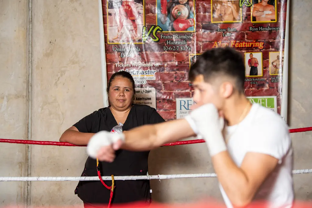 A woman leans on the ring ropes and watches as a young boxer in hand wraps throws a blurred punch in the foreground.