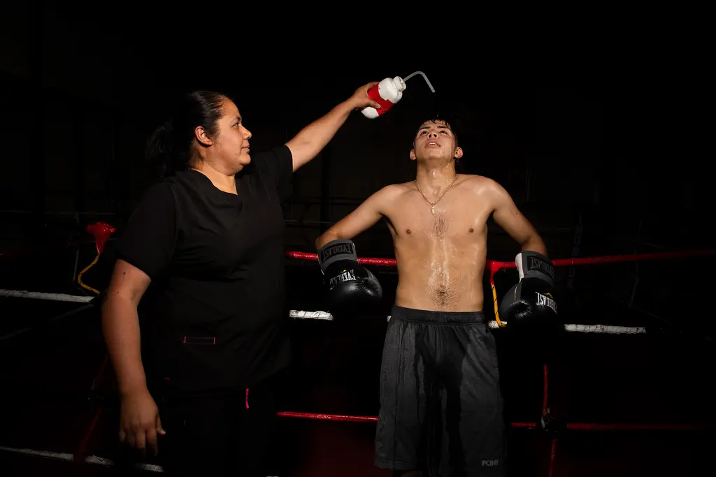 A woman squirts water from a red and white bottle over a sweating young boxer leaning on the ropes in a dark ring.