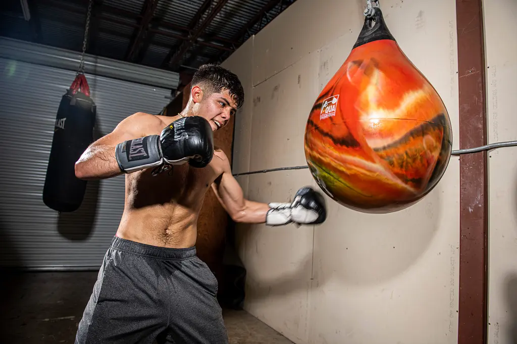 A young boxer in black gloves throws a hook into an orange water bag beside a gym wall.