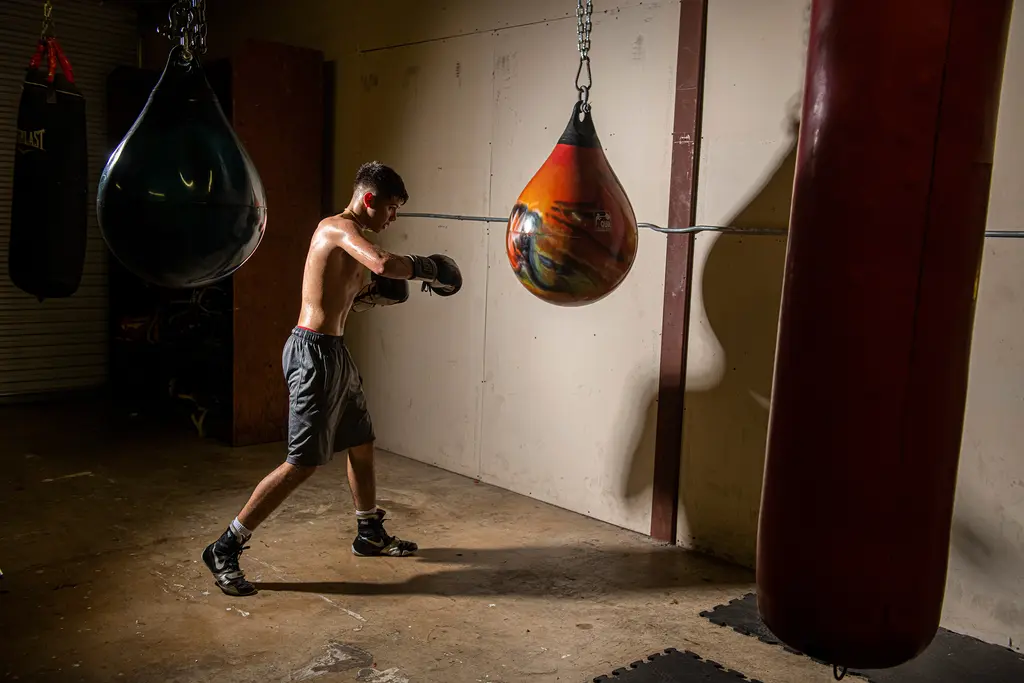 A young boxer throws punches at an orange water bag in a dim gym lined with heavy bags.