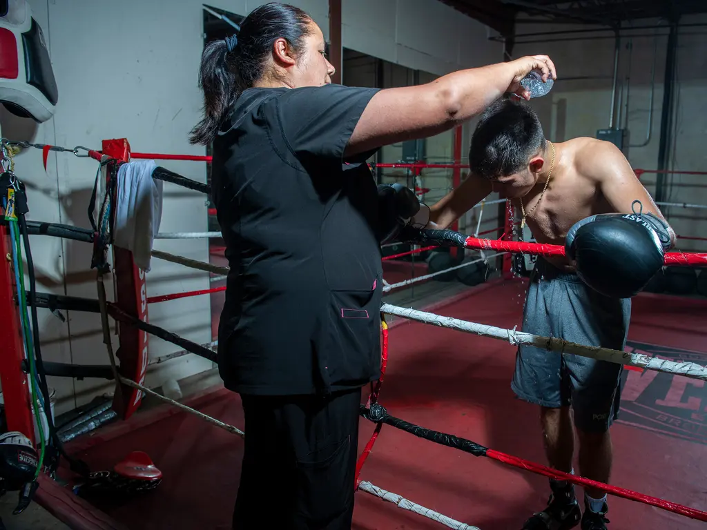 A woman pours water from a bottle over the head of a young boxer bent over the ring ropes.