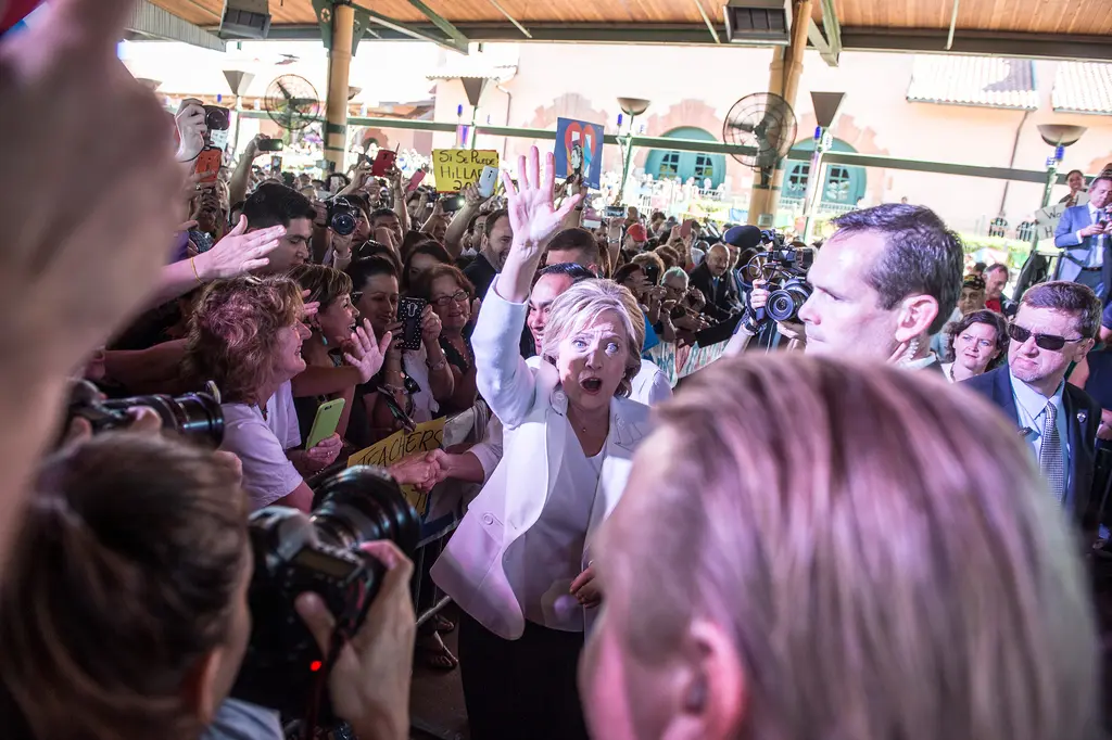 Hillary Clinton, in a white jacket, waves and reaches toward a crowd of supporters as photographers crowd the rope line.