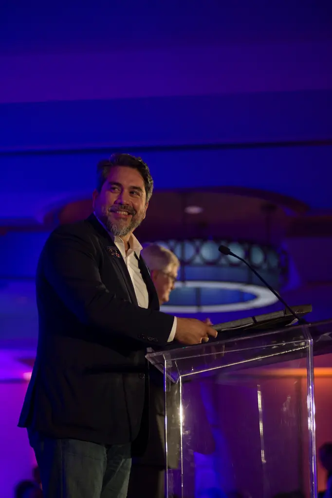 A bearded man in a dark blazer and open-collar shirt stands at a clear lectern under blue light, smiling toward the audience.