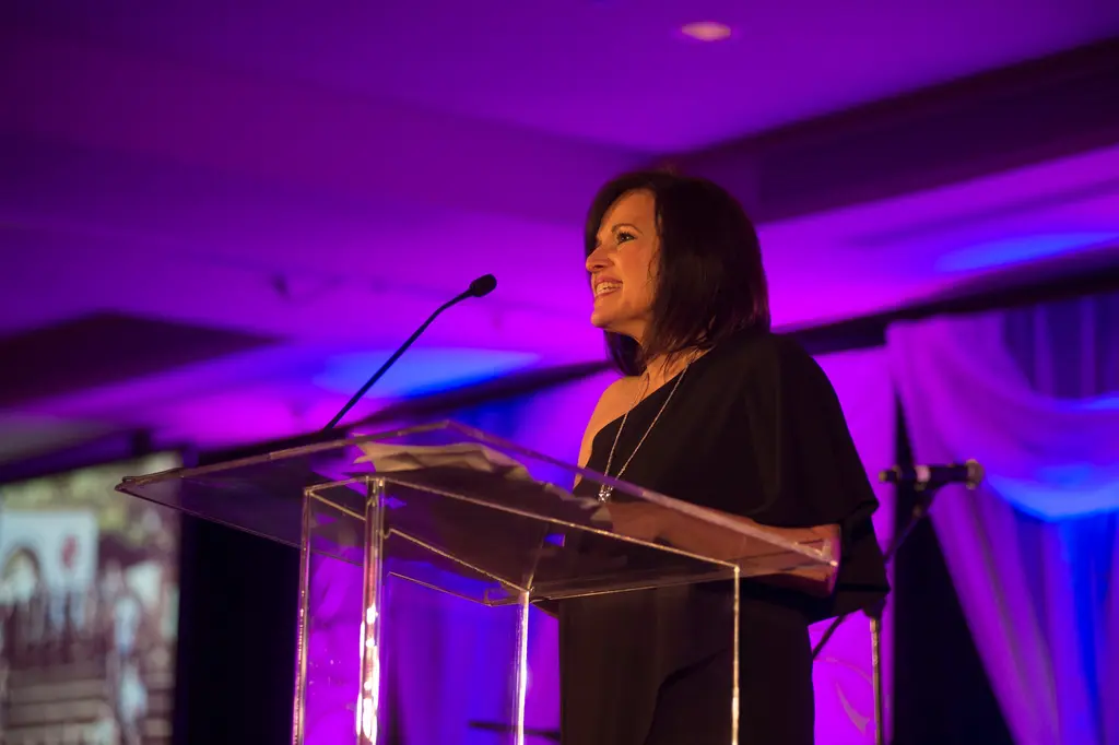A woman in a black dress stands at a clear podium under purple light, smiling as she speaks.