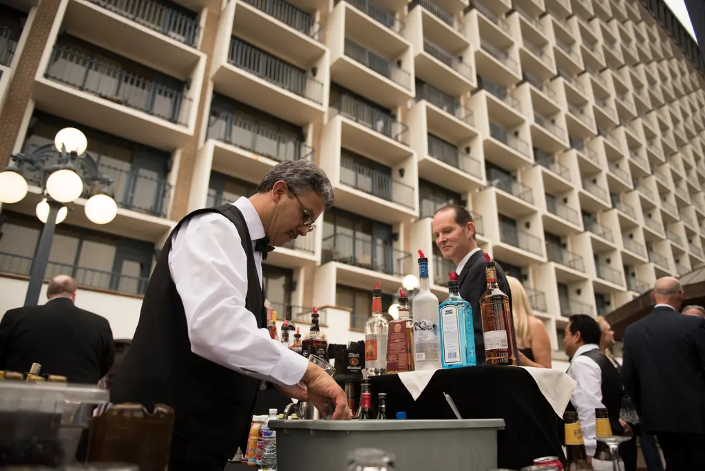 A bartender in a black vest and bow tie works at an outdoor bar lined with bottles, under the rows of balconies on the hotel tower.